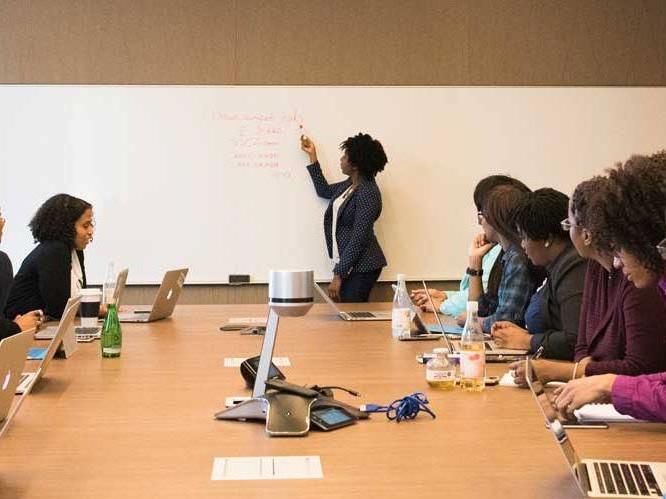 Woman is pointing to the white board, talking to the group of students who are focusing on her